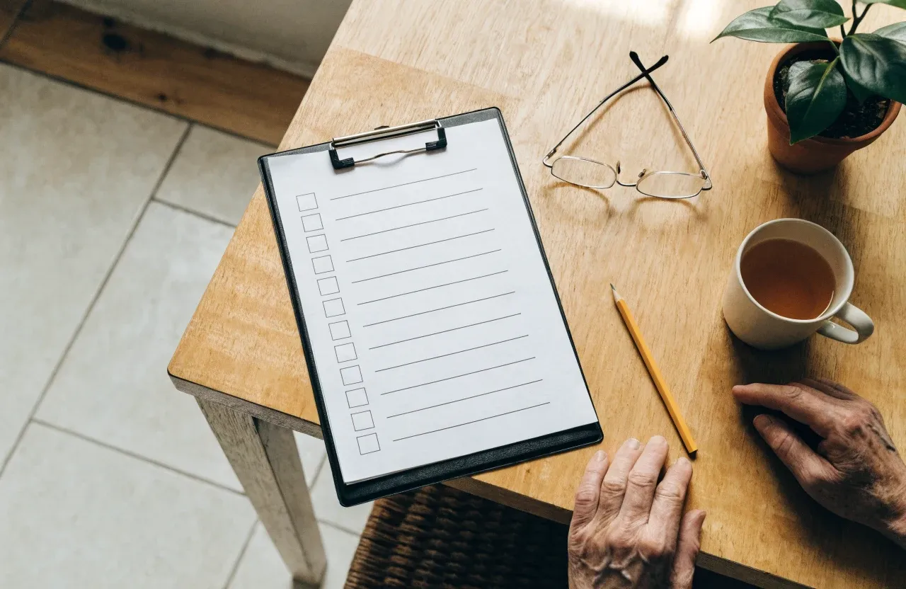 A checklist on a clipboard on a kitchen table, with reading glasses, a pencil and a mug of tea