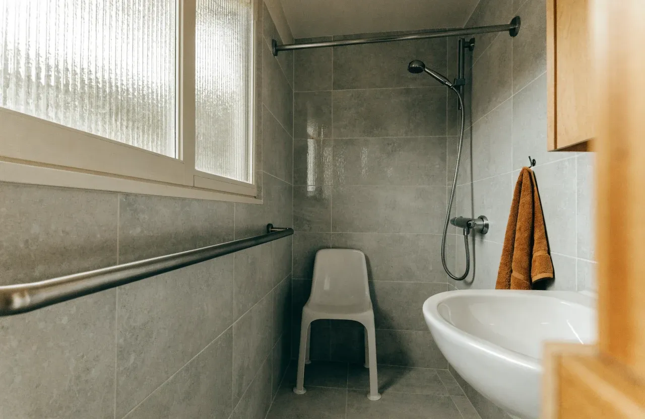 A tiled walk-in shower with a stainless-steel grab bar and a white shower chair